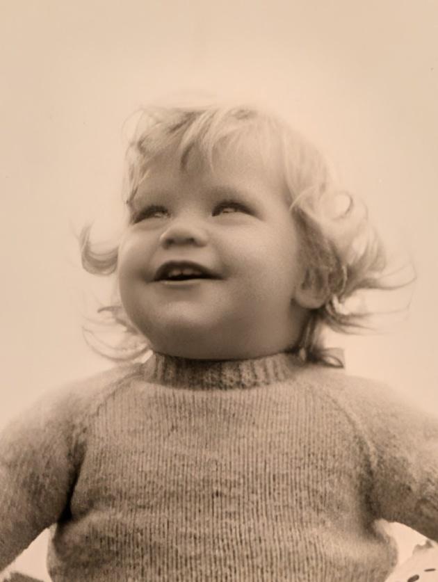 Nicky as a fair-haired toddler in a knitted jumper, laughing and looking upwards
