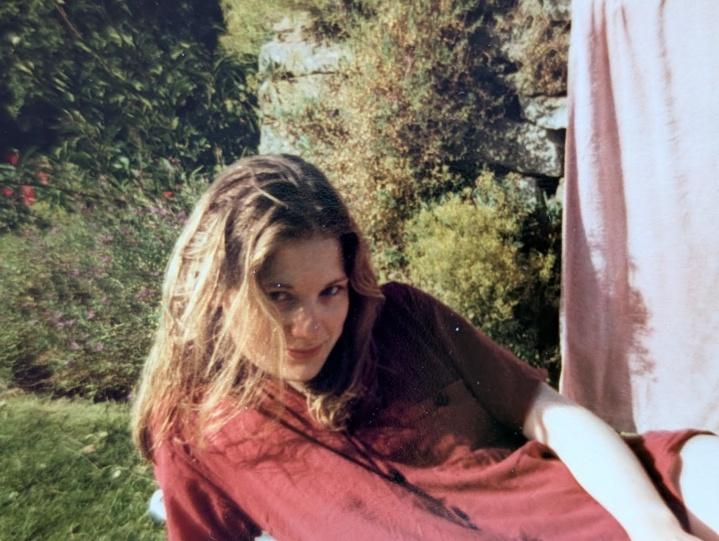 Nicky as a young woman with long hair, in a red top, sitting in a sunlit garden