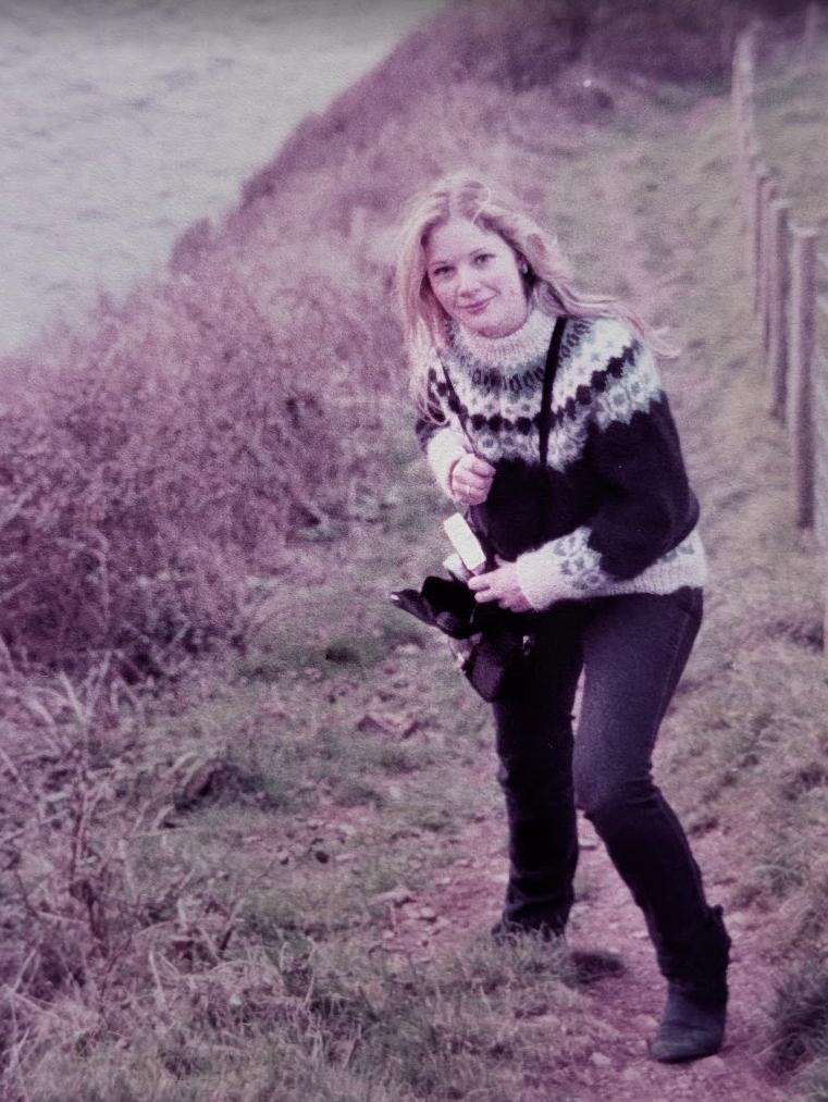 Nicky in a patterned Icelandic jumper on a grassy coastal path, turning to smile at the camera