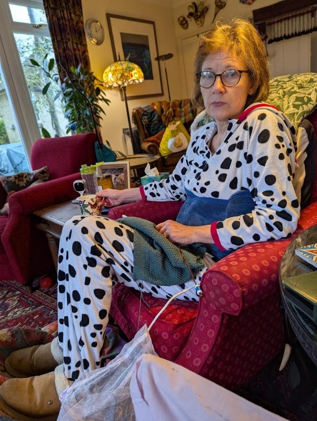 Nicky knitting in an armchair at home, wearing dalmatian-print pyjamas, a cup of tea beside her