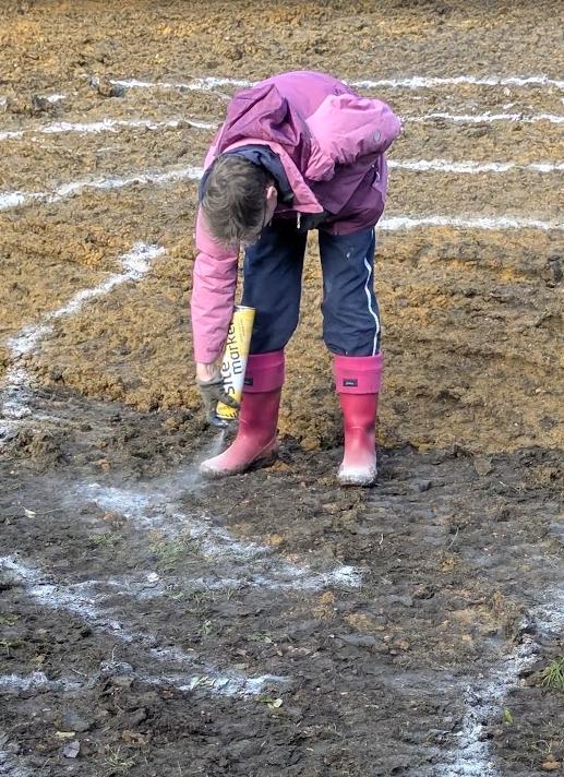 Nicky in a pink raincoat and wellington boots, bending down to spray-paint the outline of the pond onto bare earth