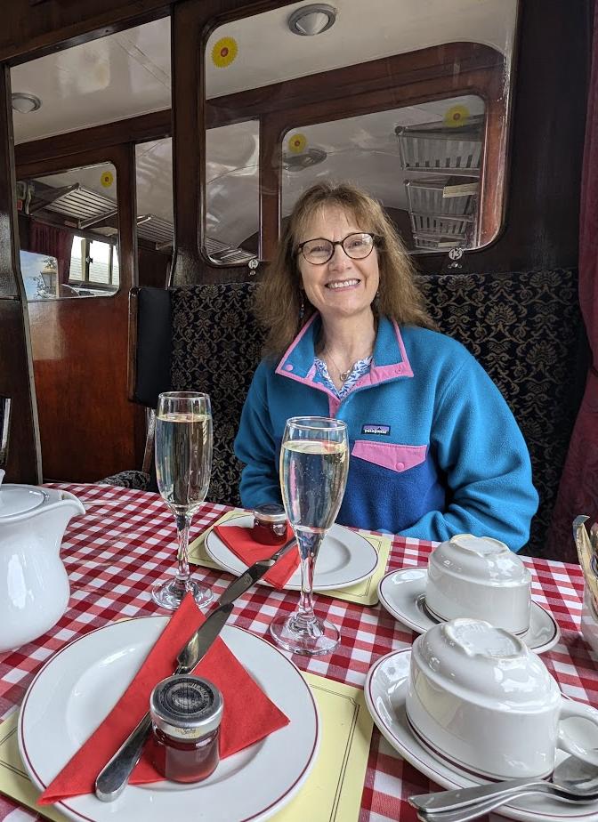 Nicky at a red-checked table in a vintage railway carriage, smiling behind two glasses of fizz