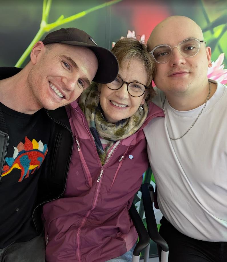 Nicky in a pink raincoat and scarf, smiling between two of her sons, all three leaning together